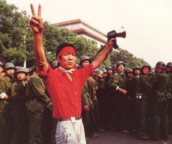 A student pro-democracy protester flashes victory signs to the crowd on June 3, as troops withdraw on the west side of the Great Hall of the People near Tiananmen Square. (Image: Public Domain)