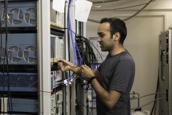 PhD student Yadav Kandel uses an Arbitrary Waveform Generator to manipulate qubits. // Assistant professor of physics John M. Nichol is photographed in his lab in Bausch & Lomb Hall along with PhD students Yadav Kandel and Haifeng Qiao September 10, 2019. The group has a paper that will be published in Nature later this month. Nichol's research brings scientists one step closer to creating a fully functional quantum computer, a type of computer that operates on the principles of quantum mechanics. Quantum computers will be able to perform complex calculations, factor extremely large numbers, and simulate the behaviors of atoms and particles at levels that classical computers (like the laptops and desktop computers we use in our everyday lives) cannot. This could lead to new energy sources, new drug therapies, faster database searches, and code-breaking techniques. One of the challenges to creating an efficient quantum computer is figuring out how to transfer information and correct errors within a quantum system. This is where Nichol's new research comes in. Nichol and his colleagues at Purdue University demonstrated a way to transfer the quantum state of one electron to another, without moving the electrons themselves. This is an important step in showing how information can be transmitted quantum-mechanically. // photo by J. Adam Fenster / University of Rochester