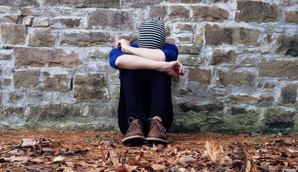 A person sits in front of a wall with knees drawn up to their chest and their head hanging down.