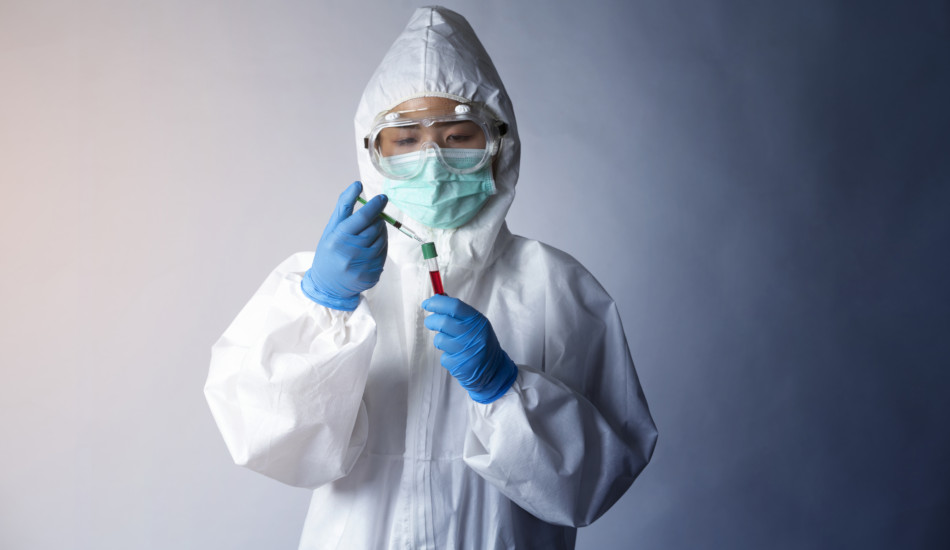 A person wearing gloves, mask, googles, and other protective clothing holds a syringe and test tube.