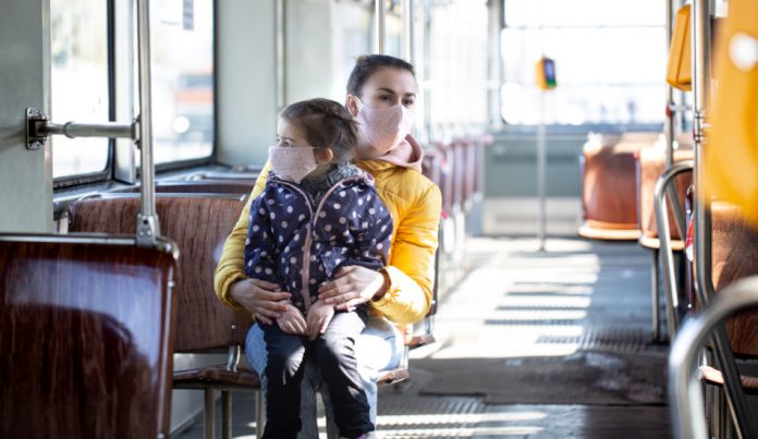 A mother with her daughter on her lap, both wearing face masks, ride on public transportation.