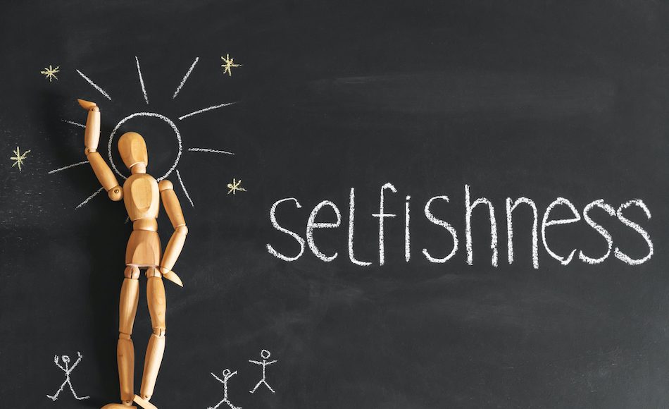 wooden figure on chalk board with white chalk written selfishness.