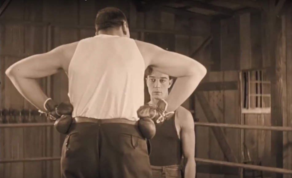 Buster Keaton's face framed by the triangle shape of a huge boxer with his back turned to camera and hands on his hips to create the triangle.