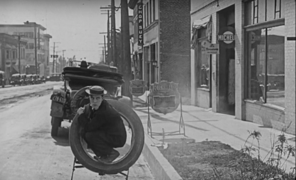 Buster Keaton crouches into a tyre that looks like it is secured to the back of an old car, the car drives away and leaves him crouching alone in a tyre looking comedic.
