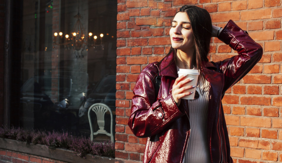 A smiling young woman in a maroon coat holds a beverage in a carry out cup while standing in front of a brick building.