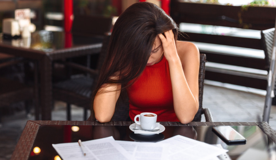 A young woman sitting at a table with a cup of coffee, some paperwork, and a cell phone while massaging her temples.