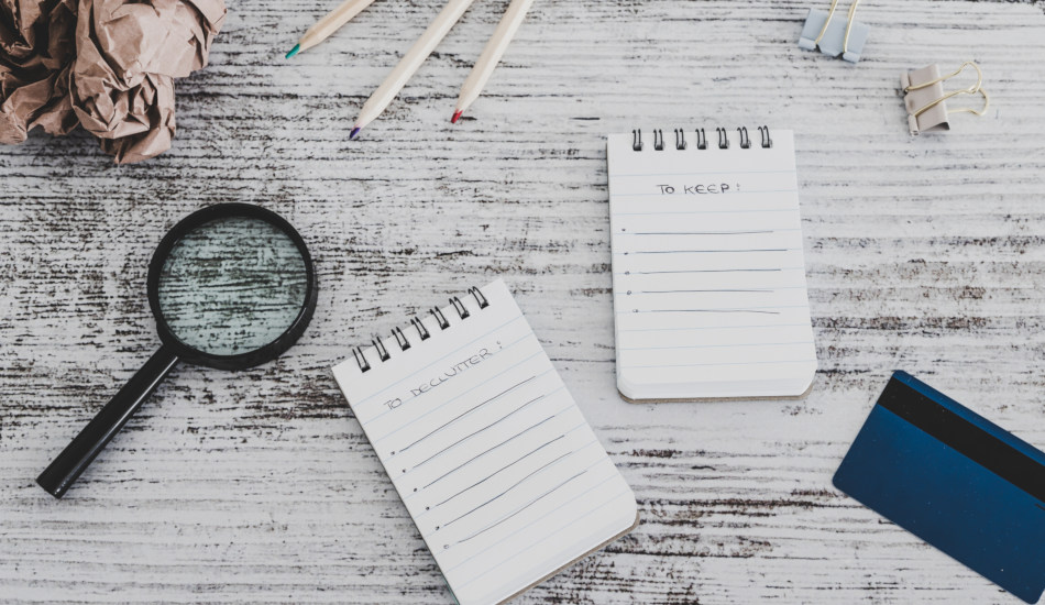 Two notebooks sit on a table with the words "To Declutter" and "To Keep" at the top, along with colored pencils, a magnifying glass, and paper clips.