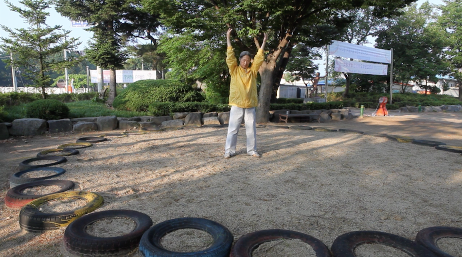 Yonghee practices the Falun Gong exercises there is a tree behind her and she wears a yellow jacket