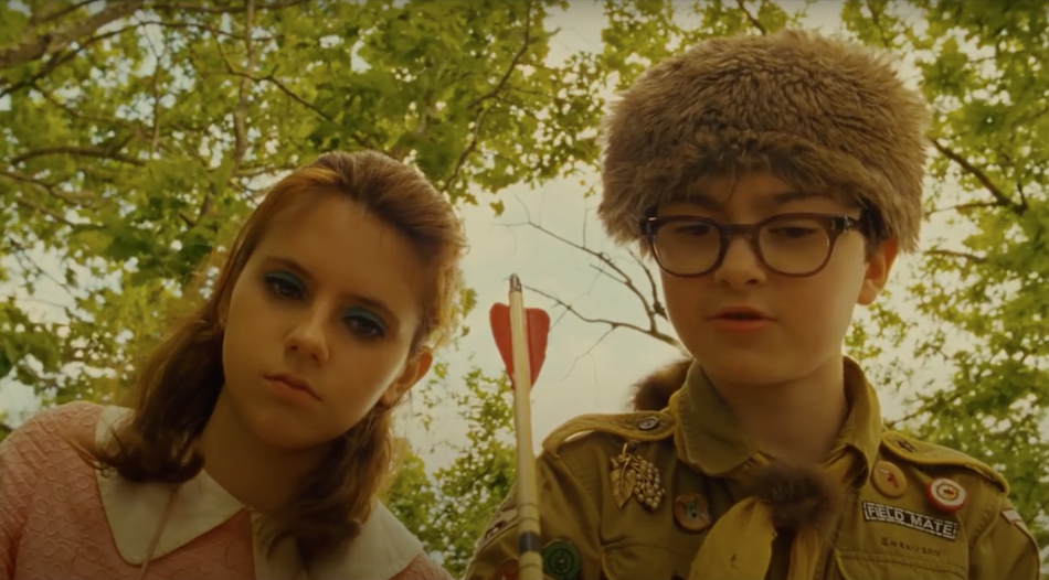 a young girl with blue eyeshadow and young boy with glasses and a scout uniform and 'davey crocket' hat hold an arrow in hand and stand above looking down