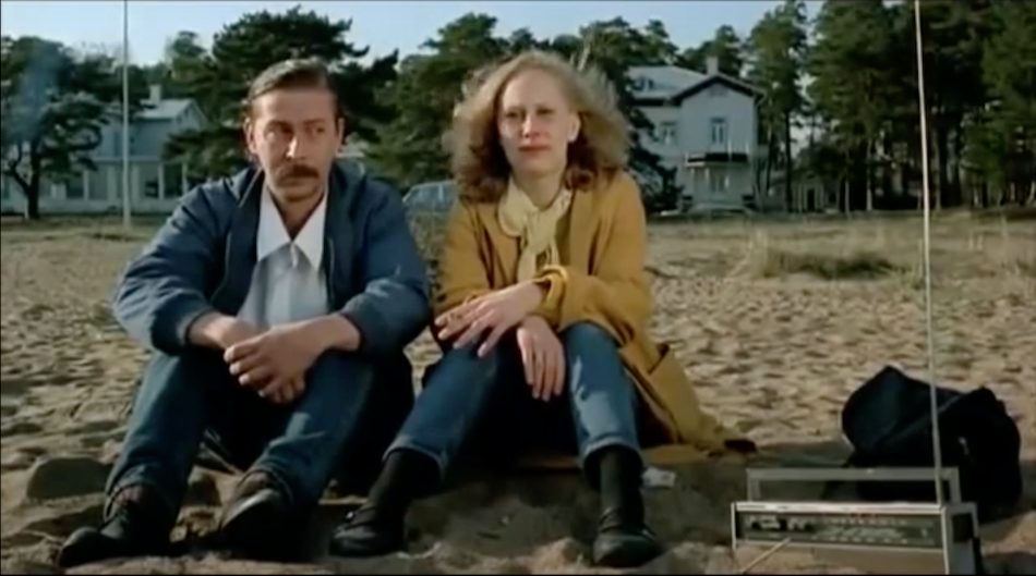 Couple sit on sand with a radio in Finland wearing jackets, jeans and boots.