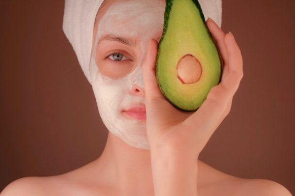 lady with hair wrapped in towel has a face mask on and is holding half an avocado over her eye