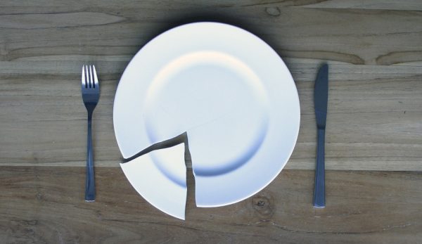 A broken plate sitting on a wooden table with a knife an fork.