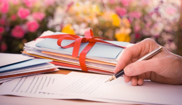 A person holding a fountain pen over a sheet of paper with a stack of evelopes tied with a red ribbon beside them on a desk with pink, yellow, and white flowers in the background.