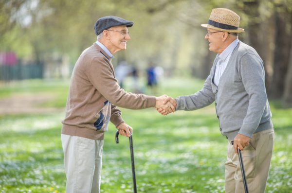 Two elderly men wearing cardigans and hats and holding canes shake hands.