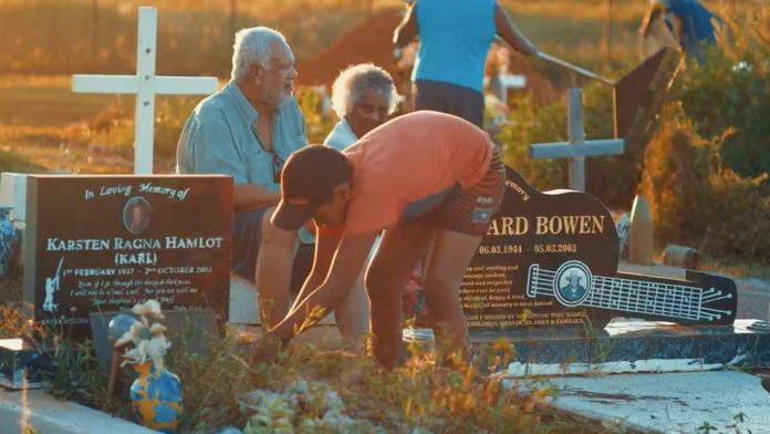 young boy weeding weeds from grave and older couple sitting in chair behind him