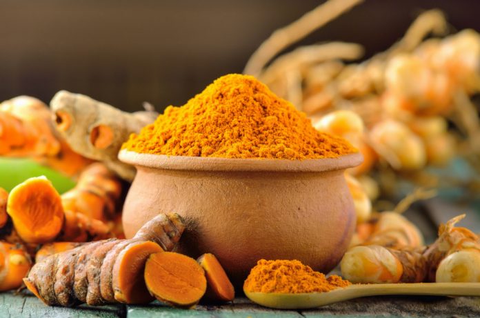 Turmeric root sitting on a table around a bowl and spoon filled with turmeric powder.