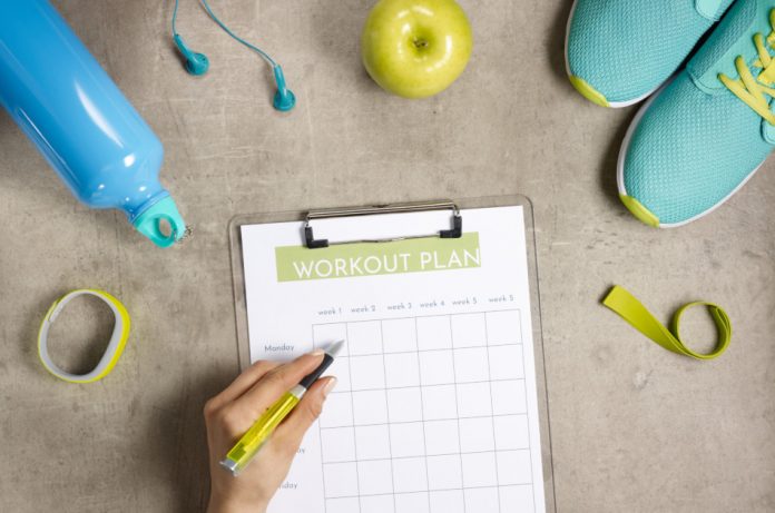 Fitness tracker, water bottle, ear buds, green apple, running shoes, and resistance band are in the picture with a woman's hand getting ready to fill out a form labeled 'Workout Plan'.