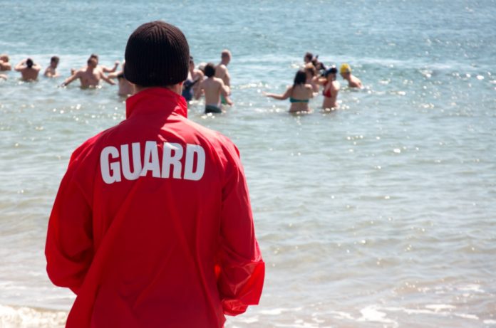 A man wearing a brown knit cap and a red jacket with the word "guard" on the back looks on as people swim in the ocean.