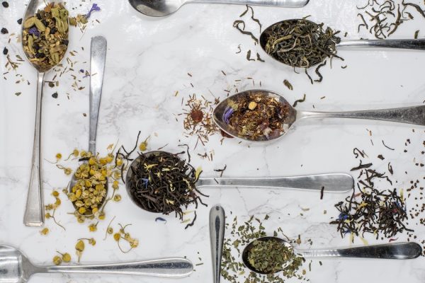 dried tea leaves of different varieties on silver spoons that lay on a white tablecloth on table around 6 spoons