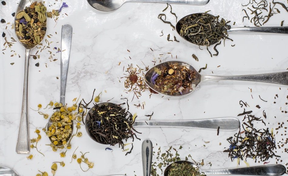 dried tea leaves of different varieties on silver spoons that lay on a white tablecloth on table around 6 spoons