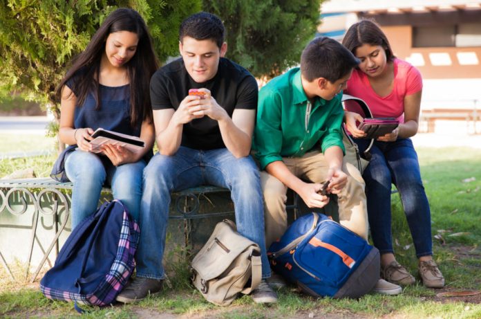 Boys and girls sit outside with school bags using mobile devices and tablets.