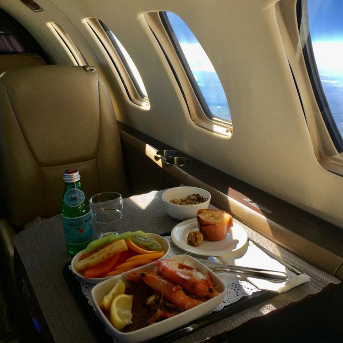 Meal on a private jet consisting of large prawns with lemon wedges, a bowl of melon, kiwi fruit and pineapple, a muffin, and a small bottle of mineral water.