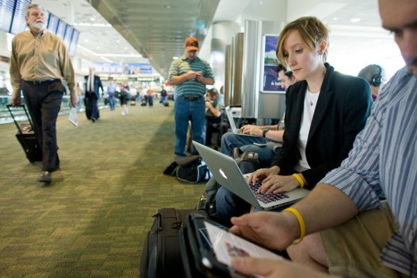 People using laptops and tablets as other passengers walk by with luggage in an airport.