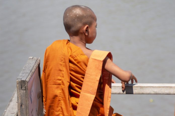 A young monk in orange robe with his back to the camera stands by a railing looking out over a lake.