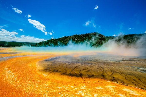 Grand Prismatic Spring with it's bright orange soil, clouds hovering and green mountain in the background, Yellowstone National Park