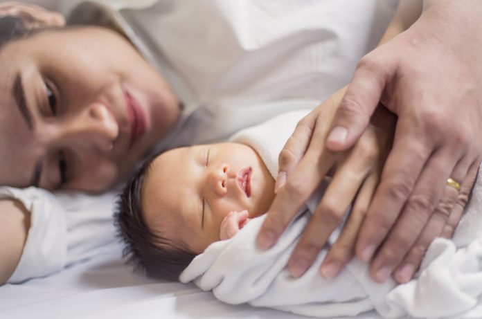 Young Asian woman lying in bed next to her baby while she and her husband each rest a hand on the child.