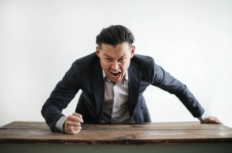 Dark haired man in a blue suit jacket leaning on a table with fist clenched and an angry expression on his face.