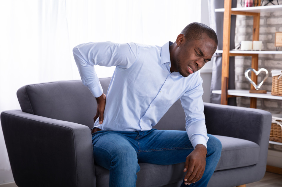 African American man holding his lower back in pain while sitting on a gray sofa.