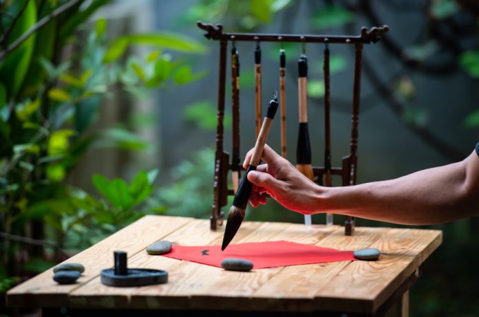 Man writing Chinese calligraphy with a brush and black ink on a red paper being held down on a wooden table by three stones.