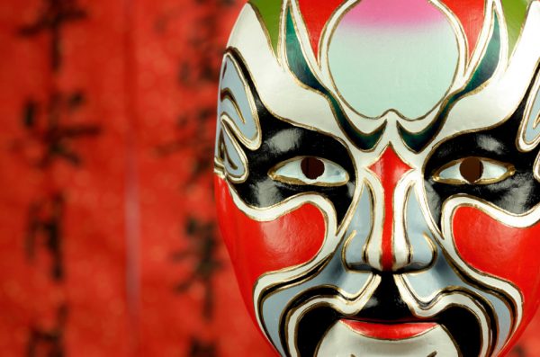 Red Chinese opera mask in front of a red background that has Chinese characters written on it in black ink.