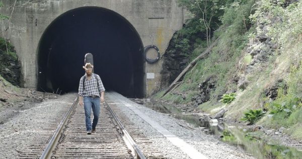 man walks down train track with guitar on back