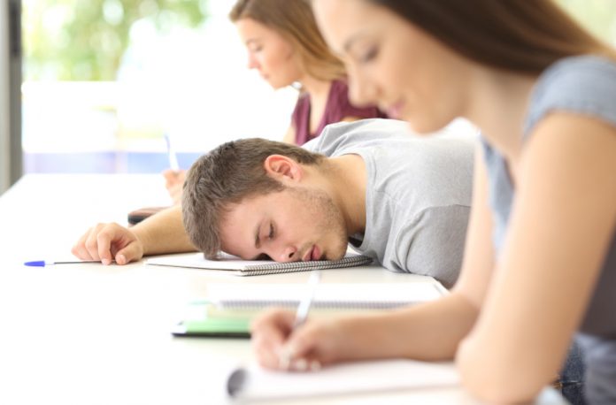 Tired male student sleeping in class between two other female classmates who are busy taking notes.