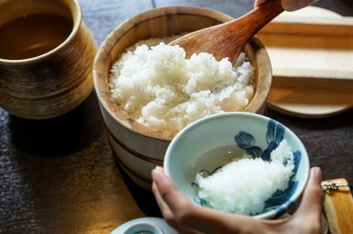 Japanese mealtime: Closeup steamed japanese white rice in traditional wooden bowl with serving hands and wooden table background, Japan.
