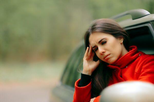 Woman in a red jacket standing by a car and rubbing her temple with a pained expression.