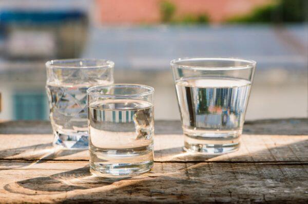 Three clear drinking glasses filled with water sit on a wooden surface outdoors.