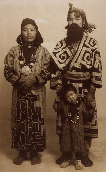 Gerhard Sisters, Public domain, via Wikimedia Commons https://commons.wikimedia.org/wiki/File:%22The_Ainu_Family.%22_(Taken_during_the_1904_World%27s_Fair).jpg An Ainu family wearing the traditional dress at a world fair in 1904. The Mother has a decorative necklace with a patterned dress with long sleeves and her lips and around her lips are tattooed in a plain colour. The child wears a jumpsuit with a stripe pattern and a beaded necklace. The father wears a dress with a pattern on it like a long coat and long pants. He has a material belt on and the father hold the toddlers hands.