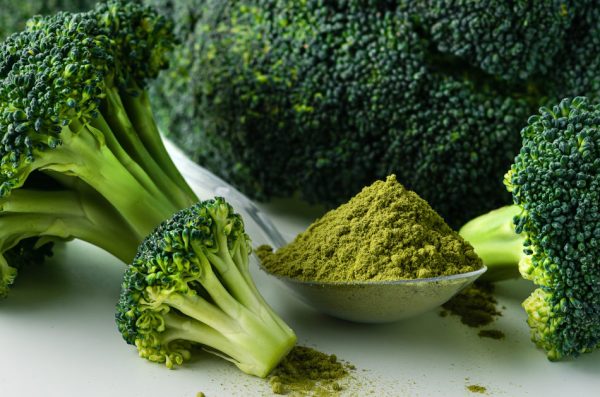 A tablespoon full of green powder sits on a white surface with a crown of broccoli.