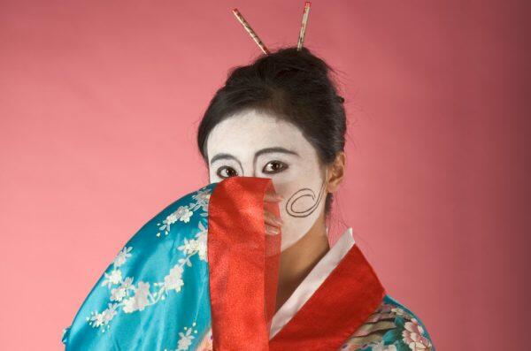 Asian female with geisha style face paint in pale blue kimono covers her mouth with one hand.