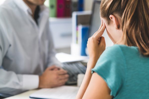 A woman rubs her temple while sitting in front of a desk while a doctor consults with her.