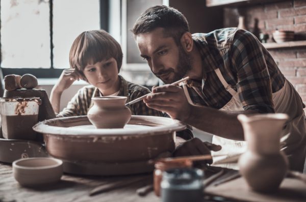 Little boy looking at confident young man drawing on ceramic pot at a pottery class.