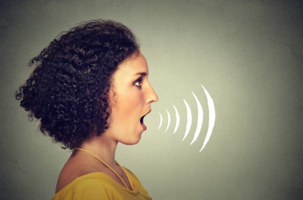 Side profile young woman talking with sound waves coming out of her mouth isolated on grey wall background.
