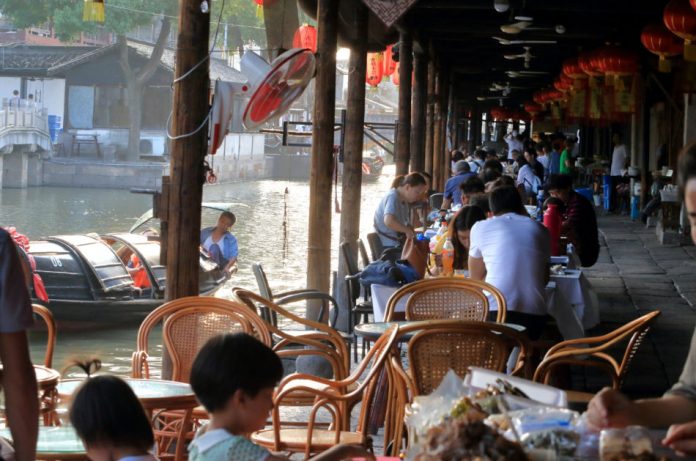 Sidewalk cafe full of customers along a waterway with a man poling a boat in Anchang, China.