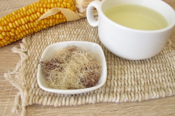 A dish filled with corn silk sits next to an ear of corn and a cup of soup.