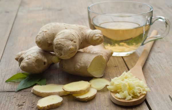Some fresh ginger sits on a wooden surface with some ginger slices, some grated ginger, a wooden spoon, a cup of ginger tea, and some mint leaves.