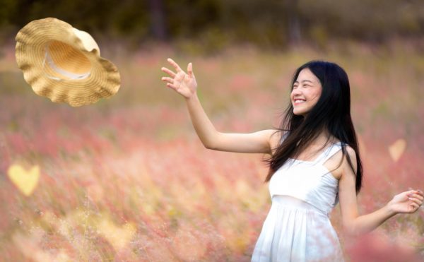 Smiling teenage Asian girl tosses her sun hat in the air while standing in a field of flowers.