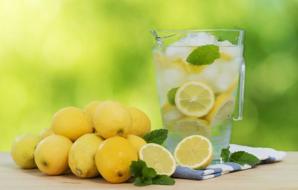 A pitcher of lemonade sits next to a stack of lemons on a wooden surface.